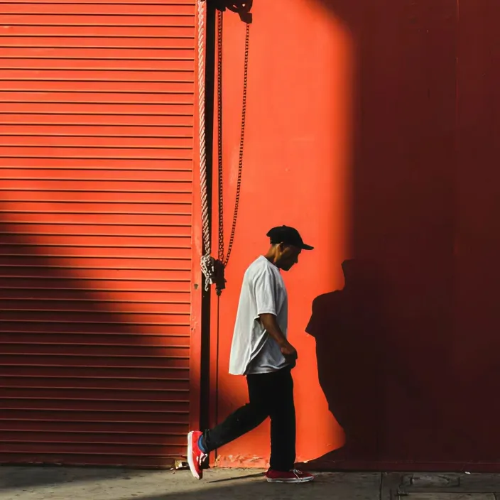 A man in a cap and white T-shirt walks past a red metal shutter and wall in low-angle light.