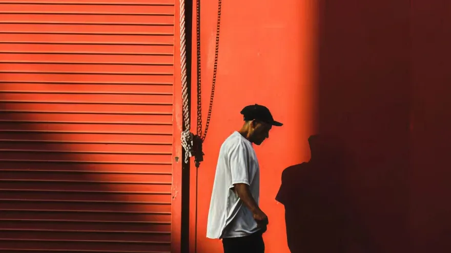 A man in a cap and white T-shirt walks past a red metal shutter and wall in low-angle light.